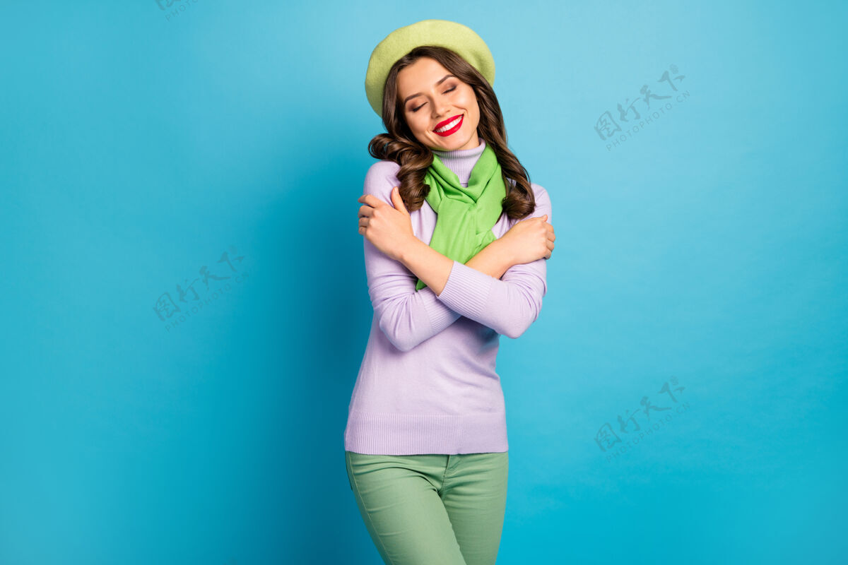 享受照片中迷人的女士闭上眼睛拥抱自己享受内心的和谐戴绿色贝雷帽紫色套头衫围巾裤子蓝色隔离墙肩膀情感帽子