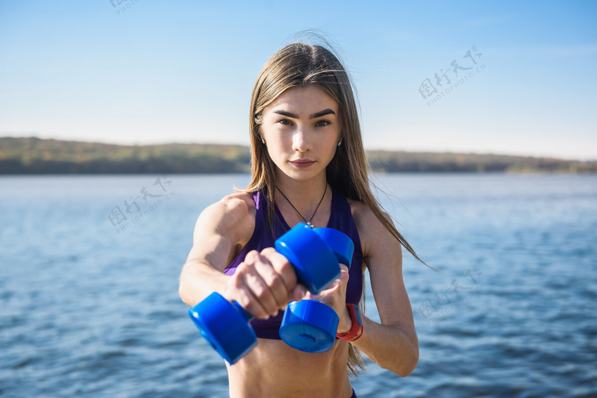 阳光美丽苗条的女人在湖边用哑铃工作健康美丽的概念锻炼哑铃衣服