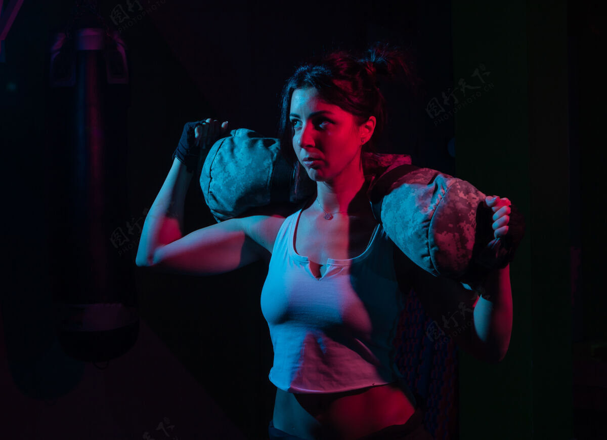 包年轻的运动女性拿着沉重的健身袋在霓虹灯梯度蓝红光下锻炼 在黑暗的墙壁上功能训练概念减肥运动员健身房