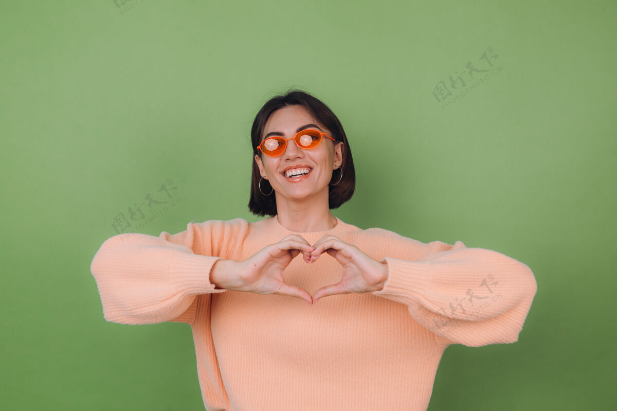 手势年轻女子穿着休闲桃色毛衣 戴着橘色眼镜 隔离在绿橄榄色的墙上 展示着用心用手爱的概念复制空间衣服年轻展示