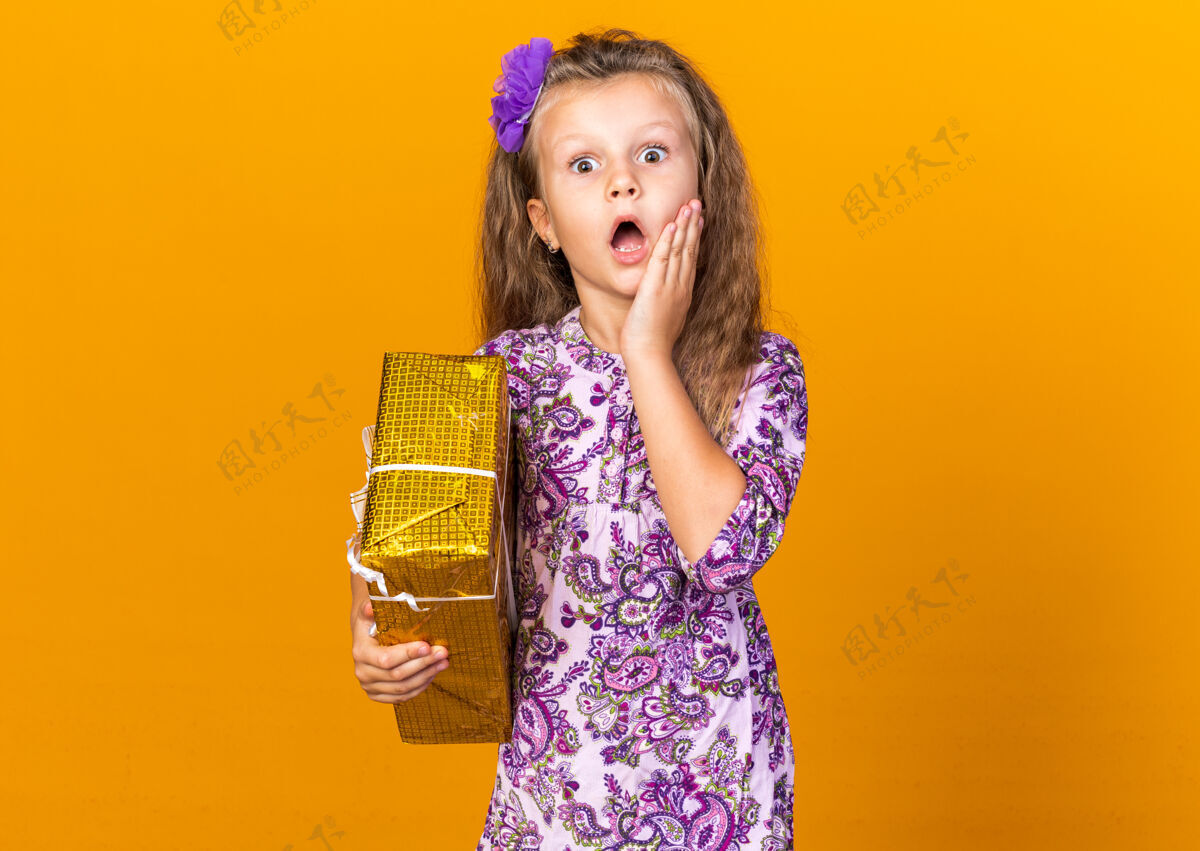 脸震惊的小金发女孩把手放在脸上 拿着礼品盒隔离在橙色墙上的复制空间抱着女孩手