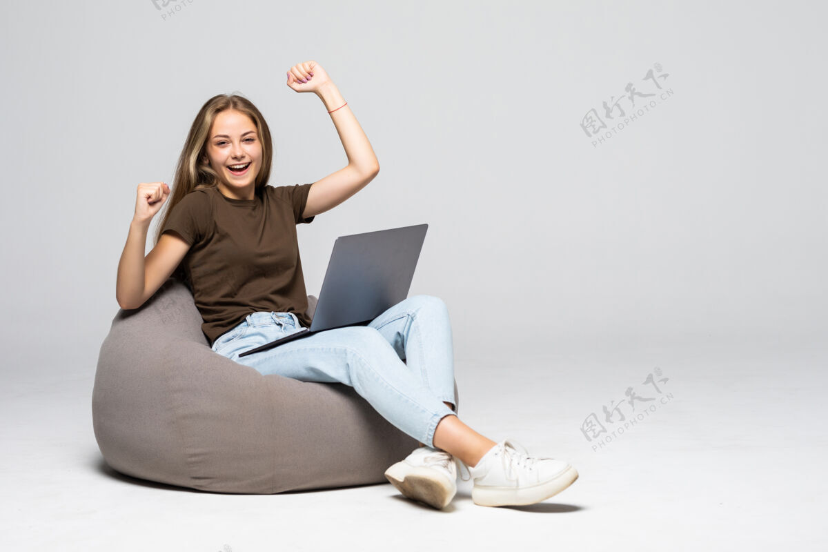微笑年轻女子坐在帕夫和笔记本电脑与胜利的姿态孤立在白墙上休闲女孩笔记本电脑