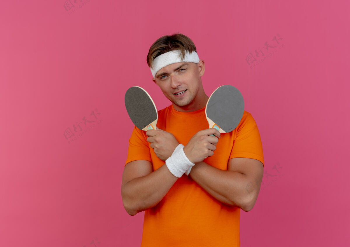 头带高兴的年轻英俊的运动男子戴着头带和手镯举行乒乓球拍隔离在粉红色的墙上运动腕带姿势