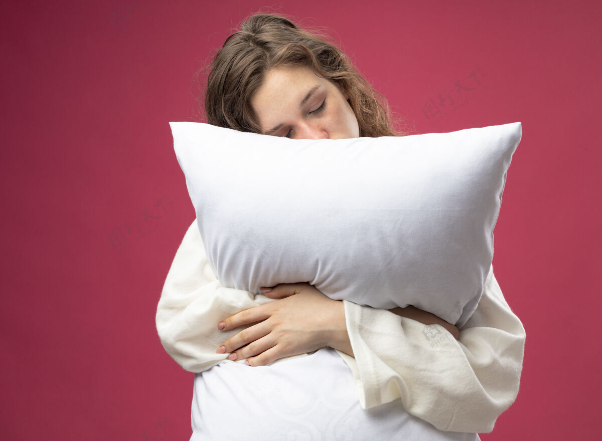 长袍悲伤的年轻病女孩闭着眼睛穿着白色长袍抱住枕头孤立在粉红色拥抱封闭眼睛