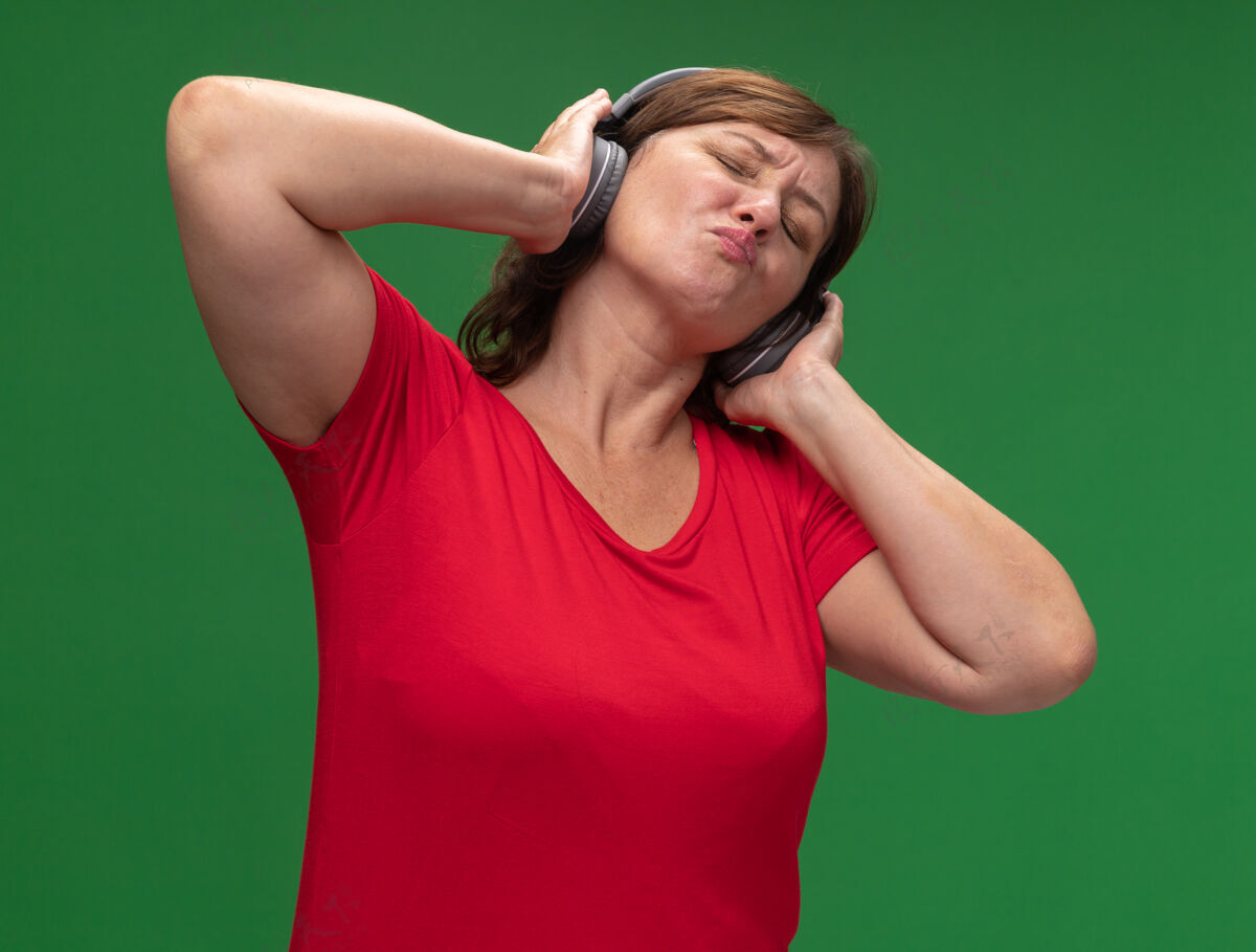 年龄快乐的中年妇女穿着红色t恤 戴着耳机 站在绿色的墙上欣赏着她最喜欢的音乐最爱耳机女人