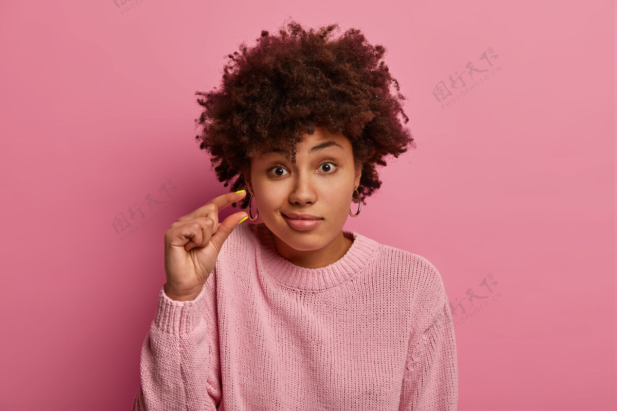 美国卷发女人的头像讨论一些很小的东西 塑造一些很小的东西 包着嘴唇 穿着休闲套头衫 孤立在粉红色的墙上 问一些小东西 做一些小动作墙小大小