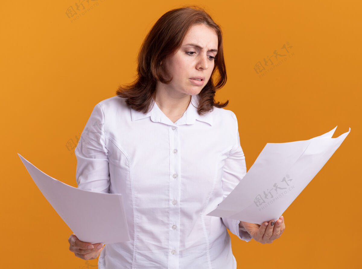 衬衫身穿白衬衫的年轻女子站在橙色的墙上 手里拿着空白的书页 困惑地看着它们空白年轻站着