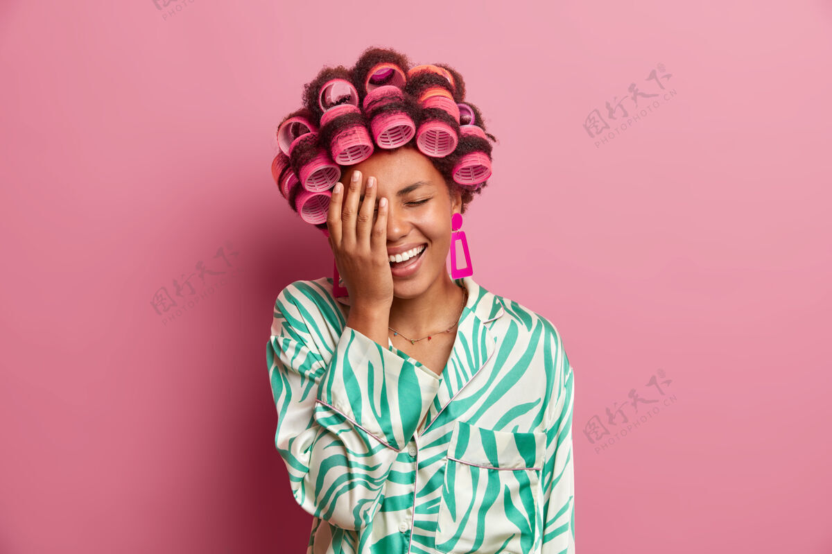 长袍搞笑快乐的女人做掌心 笑点什么 戴卷发 做完美的发型 穿睡衣 对着粉色的墙壁摆姿势女人 打扮和家居风格的概念乐观快乐肖像