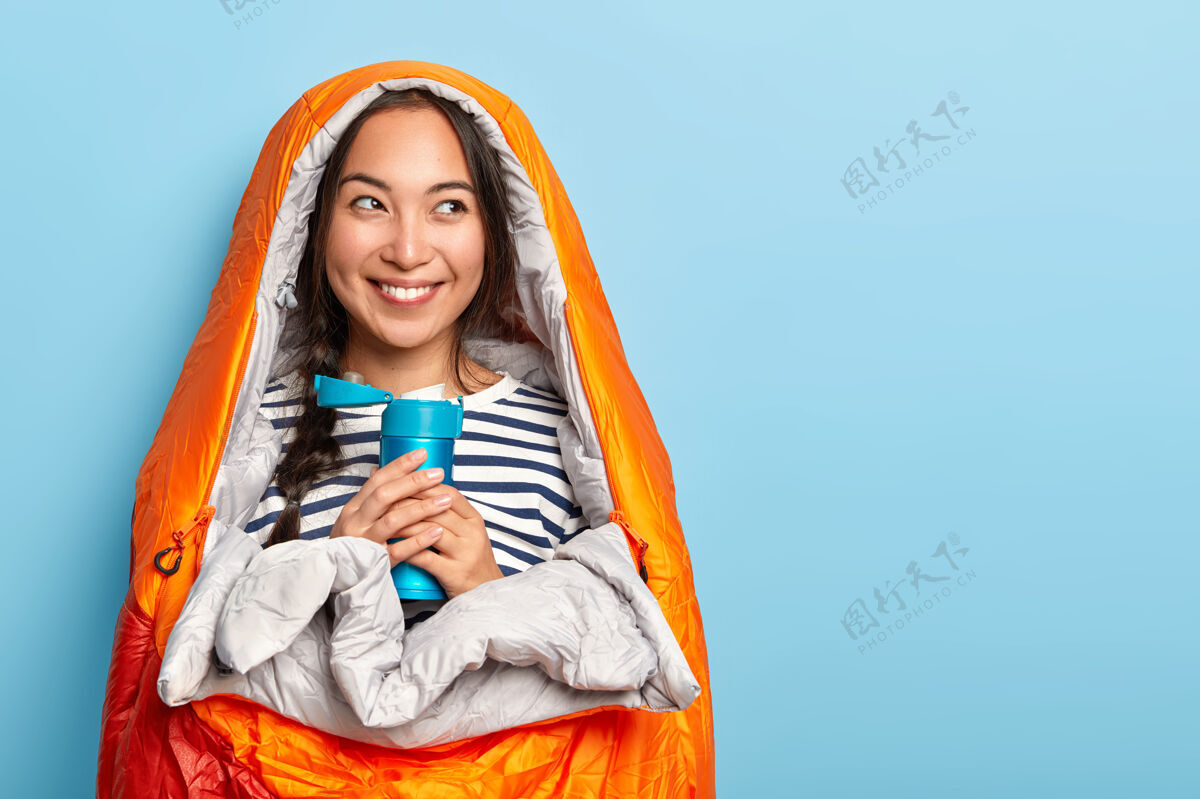 亚洲漂亮的女旅行者穿着条纹毛衣 包在睡袋里 拿着热水瓶 享受野营生活 有暑假和冒险 脸上有迷人的牙齿微笑旅行站民族
