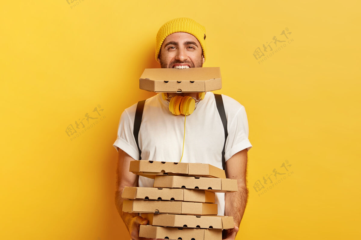 欢呼快乐男快递员超载纸箱披萨盒 手捧一叠纸箱和一个放在嘴里 穿着休闲服 隔着黄色的墙忙订单纸箱