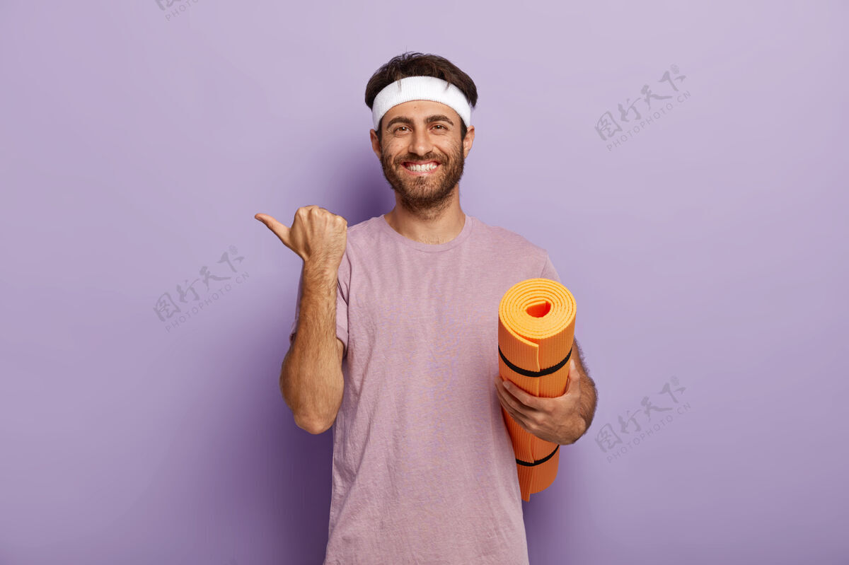 垫正面没刮胡子的家伙戴着白色头带和紫色t恤 拿着卷起的垫子 指着旁边运动头带男性