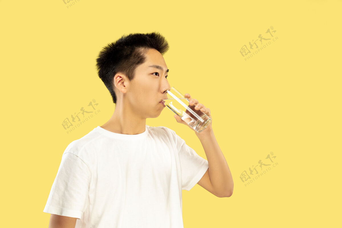 手势黄色摄影棚背景上的韩国年轻人半身像穿白衬衫的男模饮用水人类情感的概念 面部表情正面视图流行色彩男人男性工作