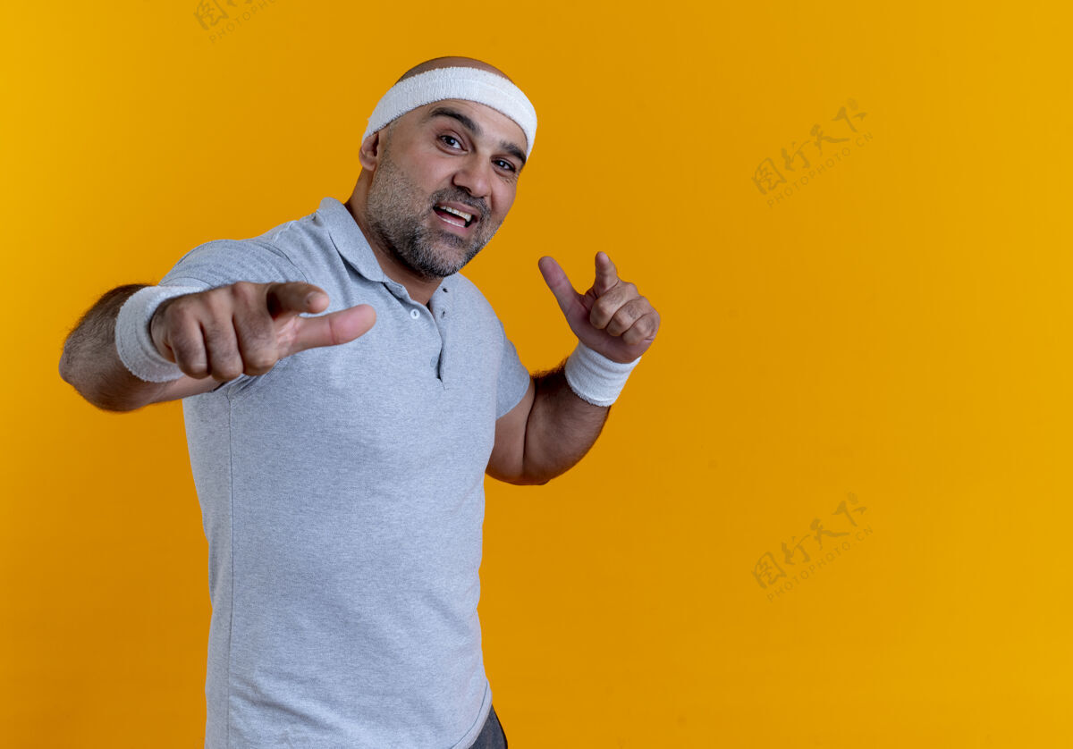 姿势戴着头巾的成熟运动型男子望着前面微笑自信地用食指指着前面站在橙色的墙上男性站着运动