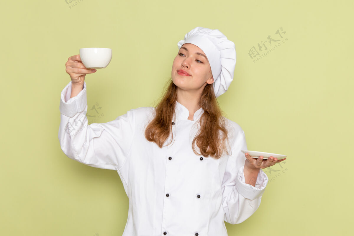 家庭主妇身着白色厨具套装的女厨师面带微笑 手持白色咖啡杯 绿色办公桌上摆放着厨房菜肴烹饪餐女颜色厨房烹饪举行