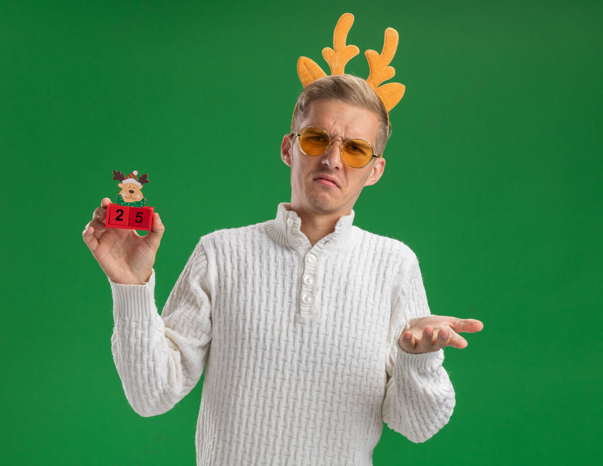 穿着皱眉的年轻帅哥戴着驯鹿鹿角头带戴着眼镜拿着圣诞树玩具和日期显示空手孤立在绿色的墙上驯鹿皱眉英俊