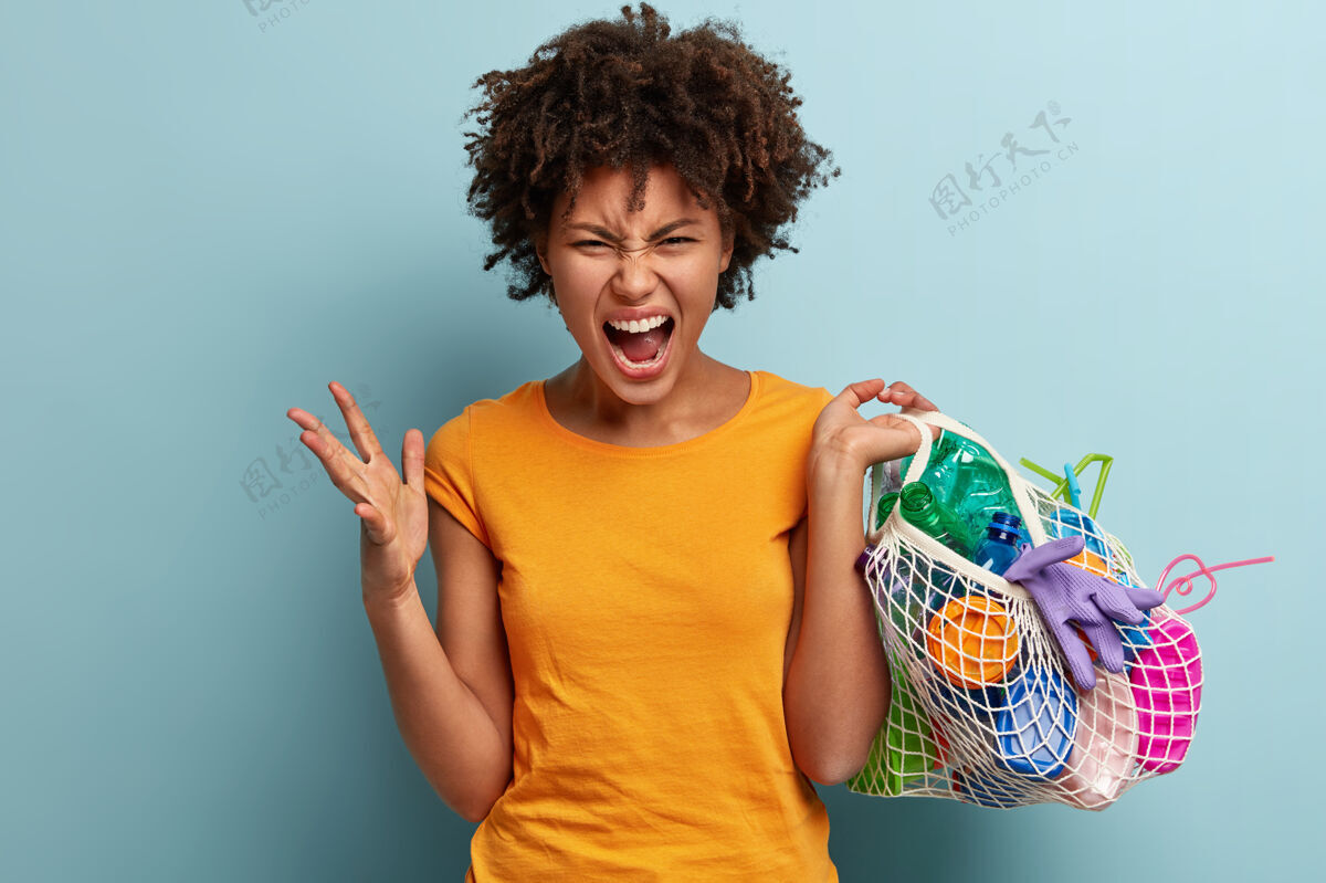 恼怒绝望愤怒的年轻女子的镜头愤怒地手势 在网袋中携带塑料物 被污染困扰 穿着橙色t恤 站在蓝色的墙壁上塑料意识概念痛苦塑料卷发