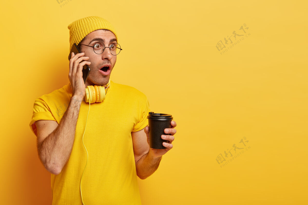 T恤惊呆了的男人有电话交谈 震惊于最新消息 穿着休闲t恤智能手机帽子饮料
