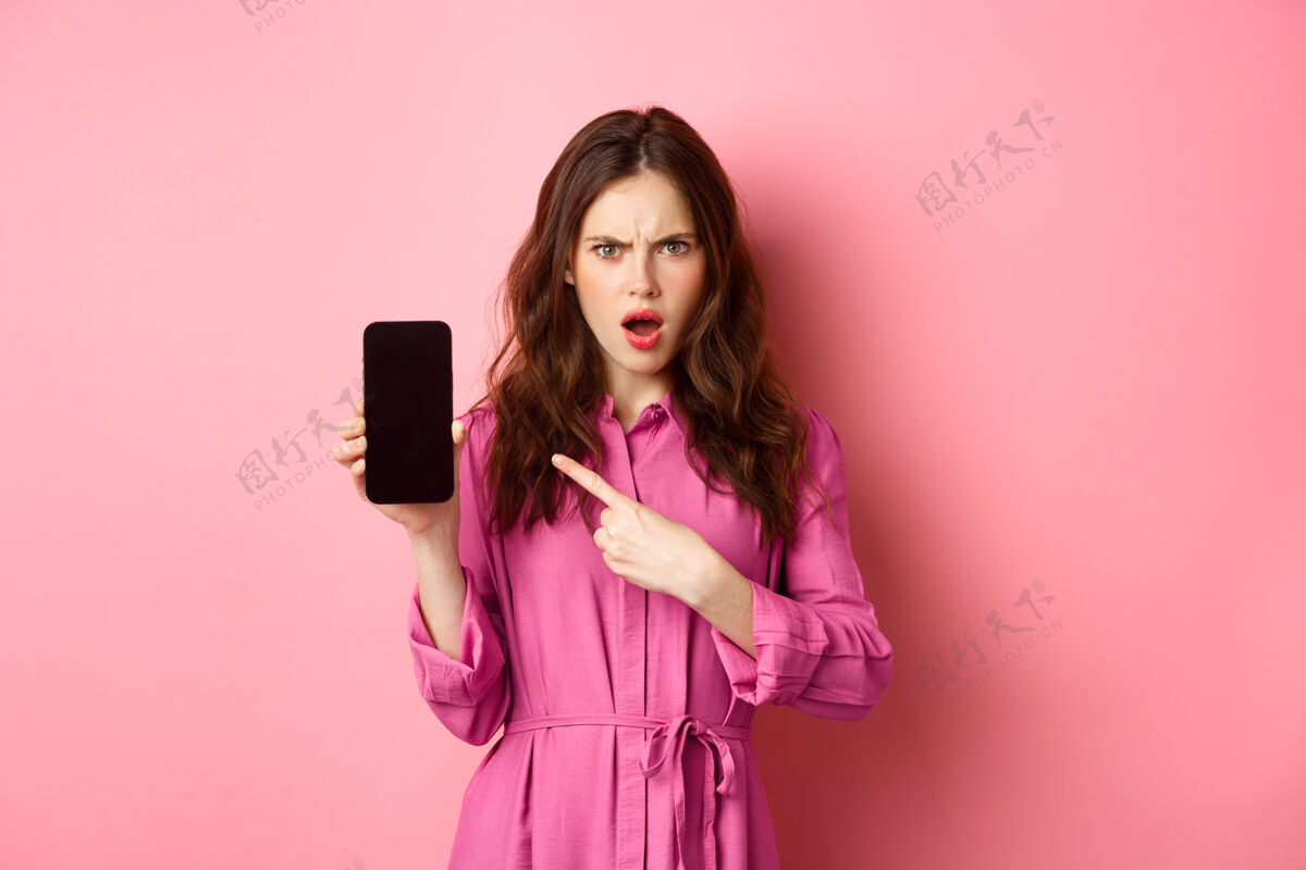 黑发技术概念愤怒的女孩指着她的智能手机屏幕 盯着相机 要求回答或解释 站在粉红色的墙上情感应用程序互联网