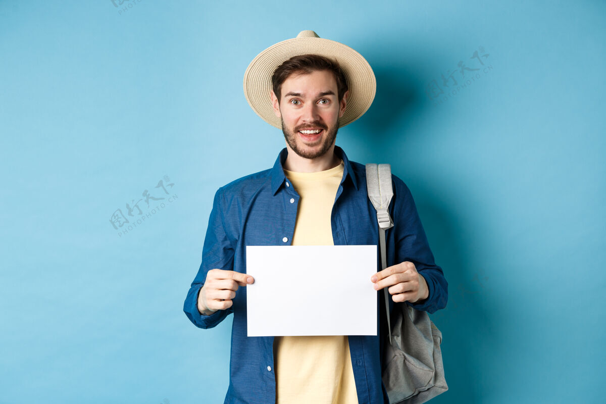 男快乐的游客戴着夏天的帽子 露出一张空纸和微笑 背着背包搭便车 站在蓝色的背景上背包朋友站