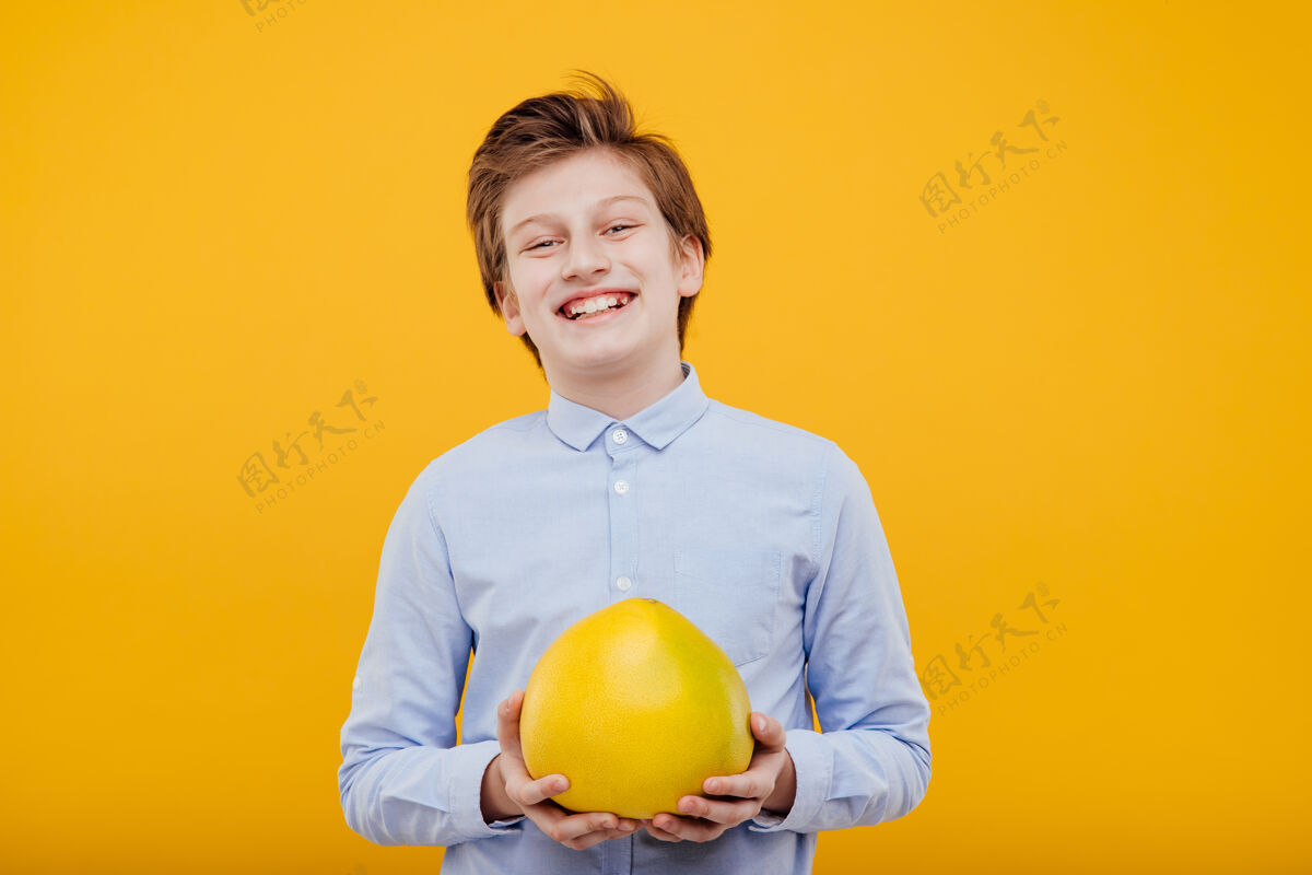 小少年快乐的男孩手里拿着柚子 穿着蓝色的衬衫 隔离在黄色的墙上肖像热带柚子水果