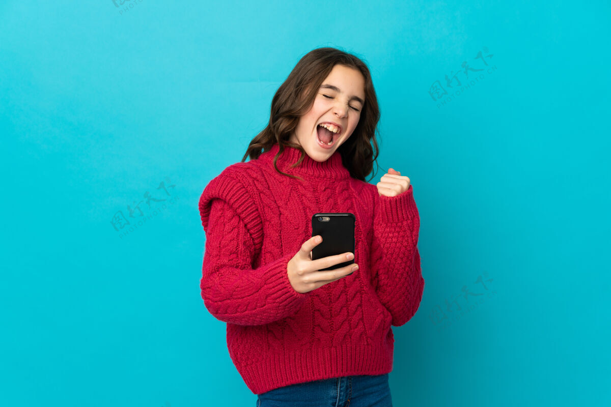 孩子小白种女孩被隔离在蓝色背景下 手机处于胜利位置女孩女性多彩