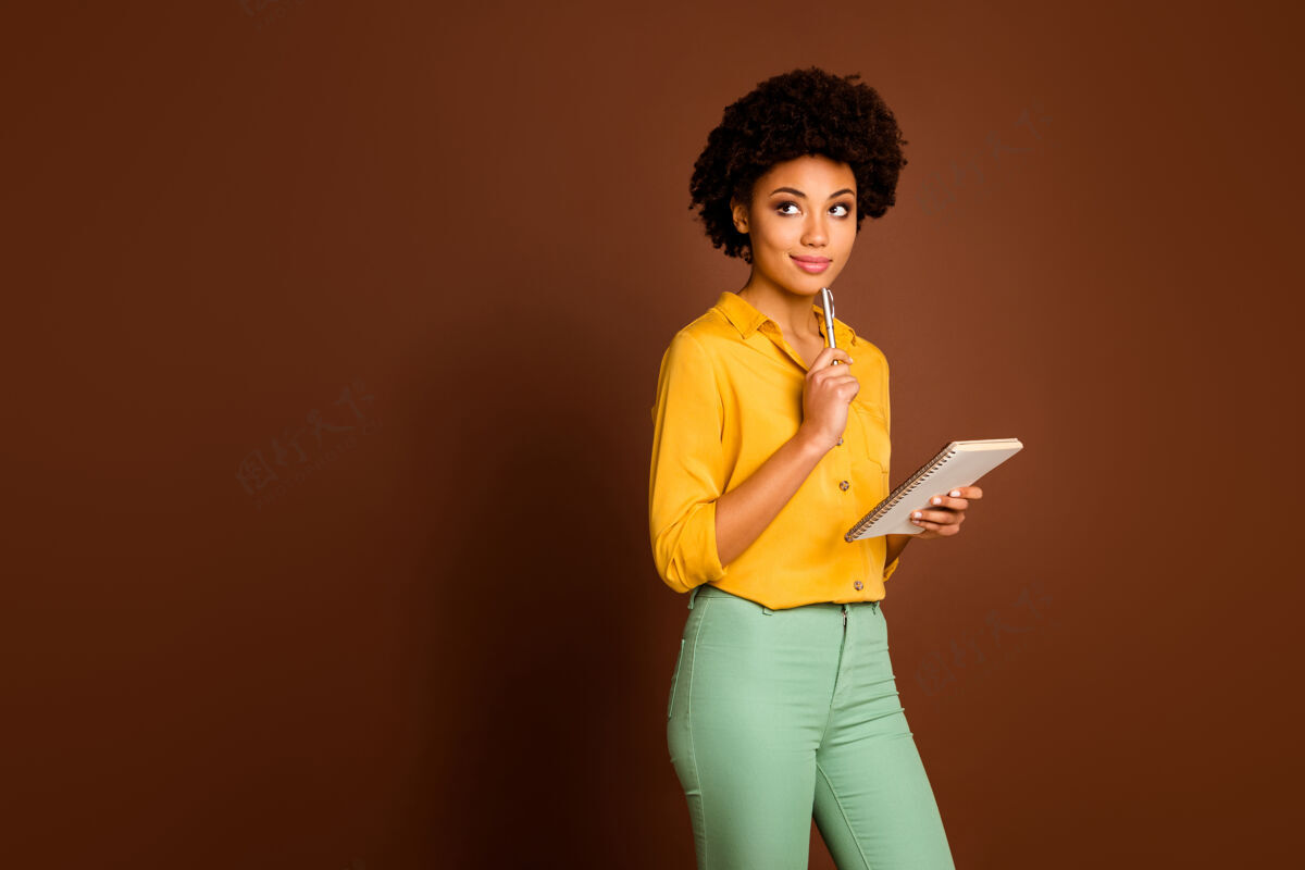 女性迷人的深色皮肤卷曲女士的照片作者拿着日记看空旷的空间等创意灵感穿黄色衬衫绿色裤子孤立的棕色非洲裔美国人灵感