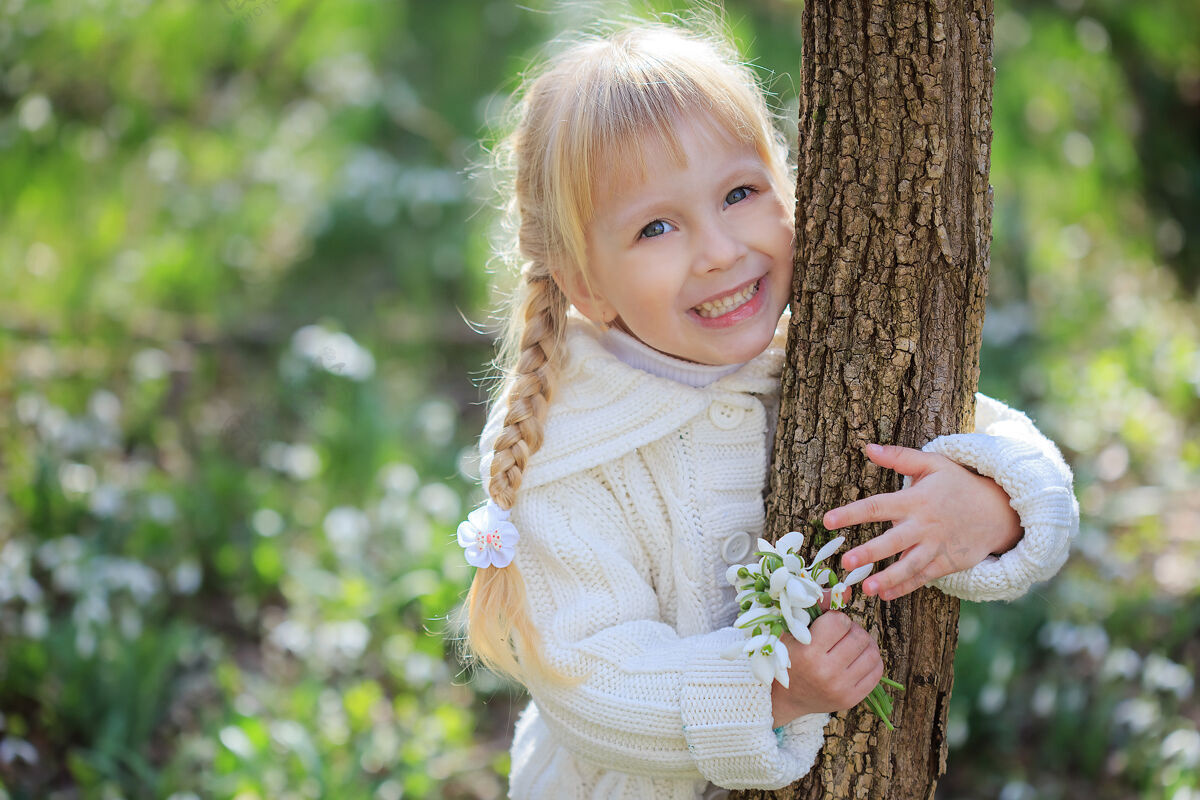 女孩带着一束雪花莲的漂亮小女孩一个穿着白色针织毛衣的小女孩抱着树干阳光明媚的春日太阳假日幼儿园