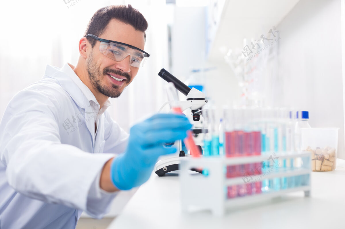 专业更多样品快乐的男同性恋实验室工作人员一边拿瓶子一边咧嘴笑 一边戴上安全眼镜研究化学家浓度