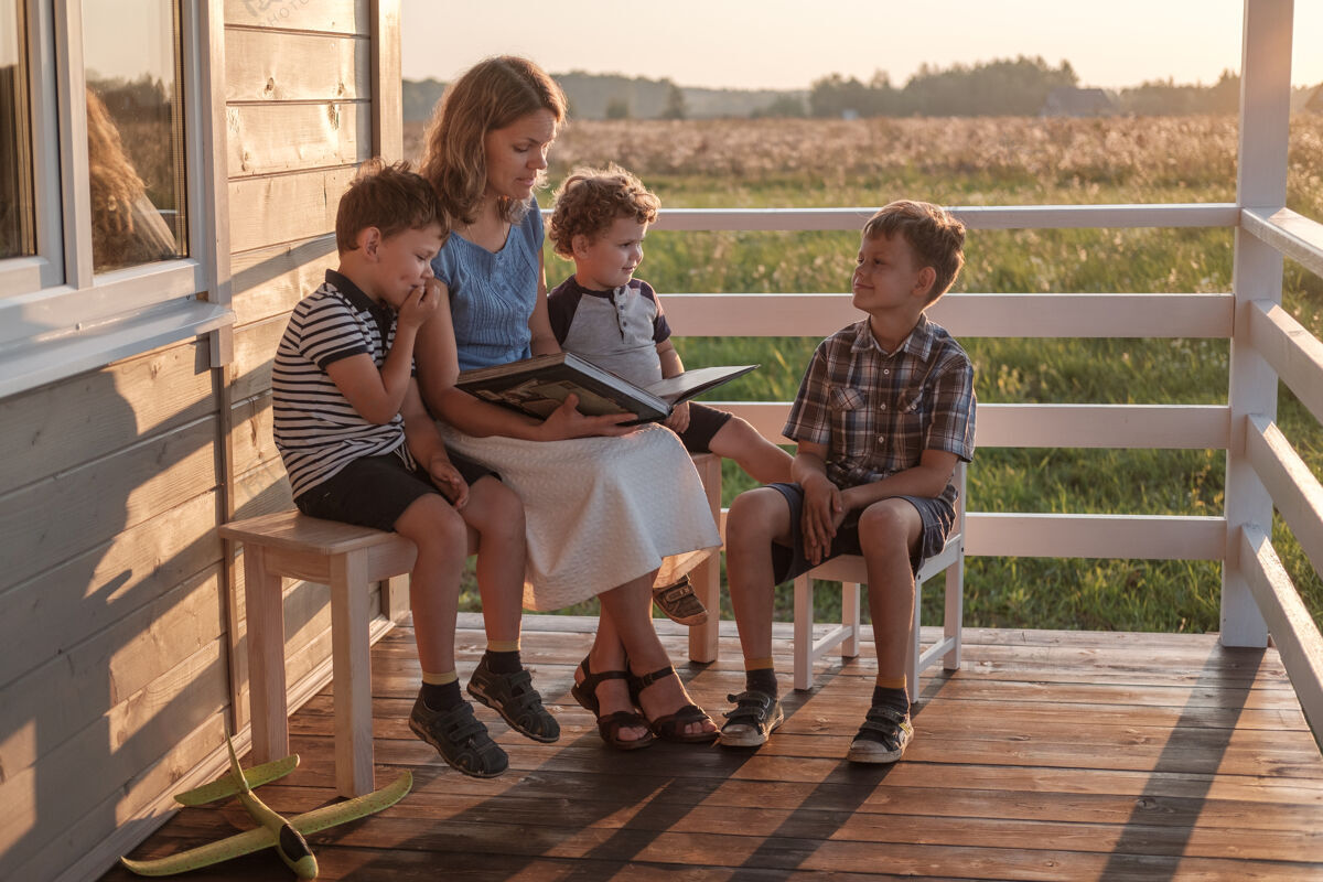 发展一位带着三个孩子的年轻母亲正在夏日阳台上看书村庄夏天白种人