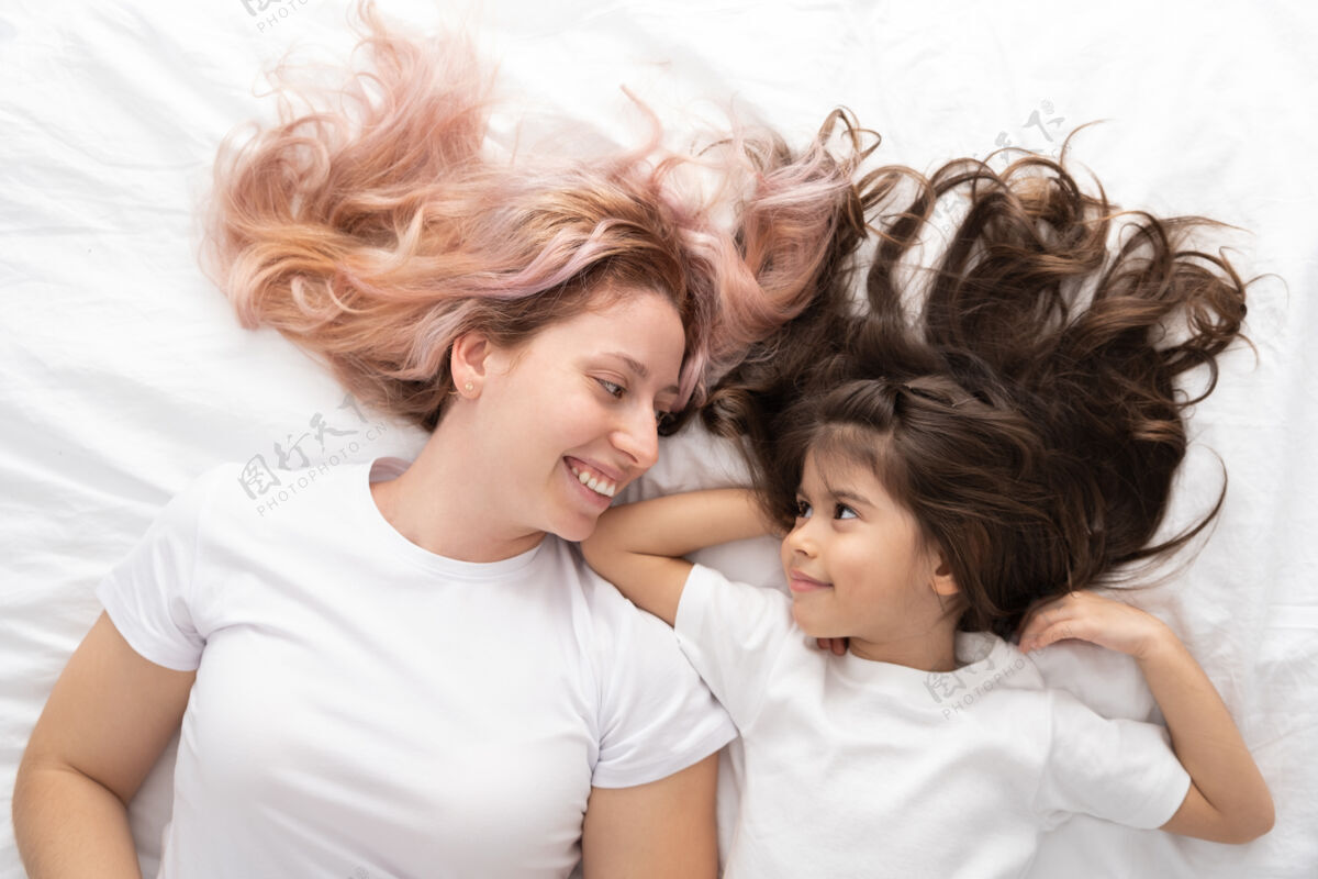 家庭年轻的妈妈和活泼的小女儿躺在家里的床上 玩得很开心童年房子母亲节
