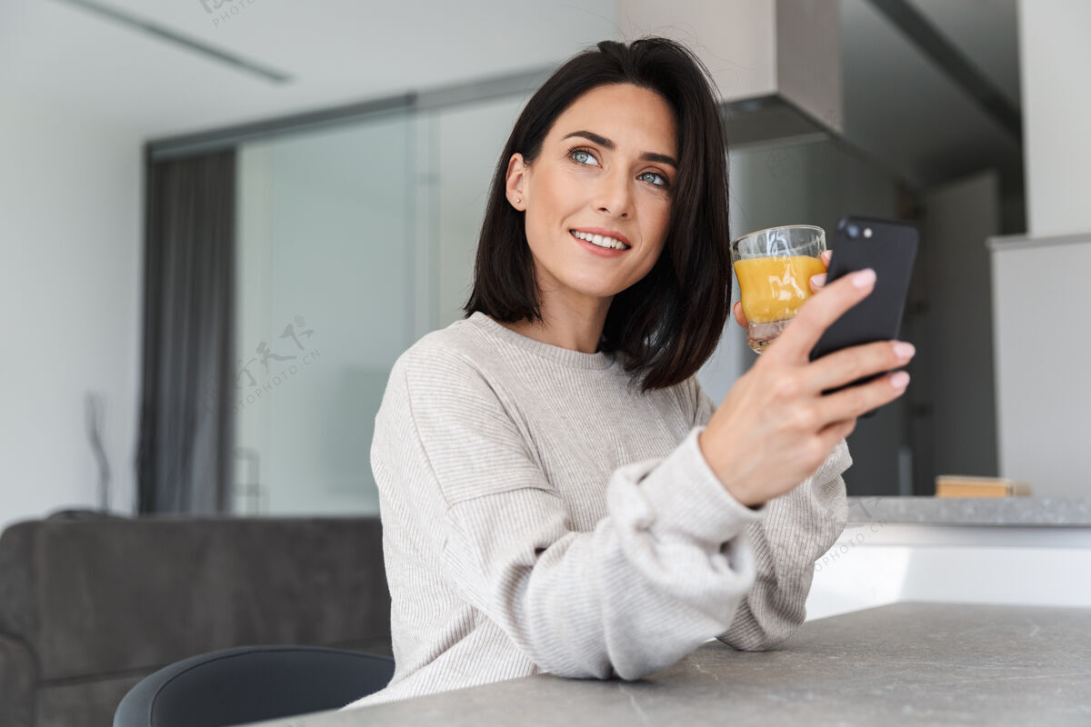 玻璃30多岁的美女一边喝着橙汁 一边用手机 一边在明亮的现代化房间里休息愉快居住沙发