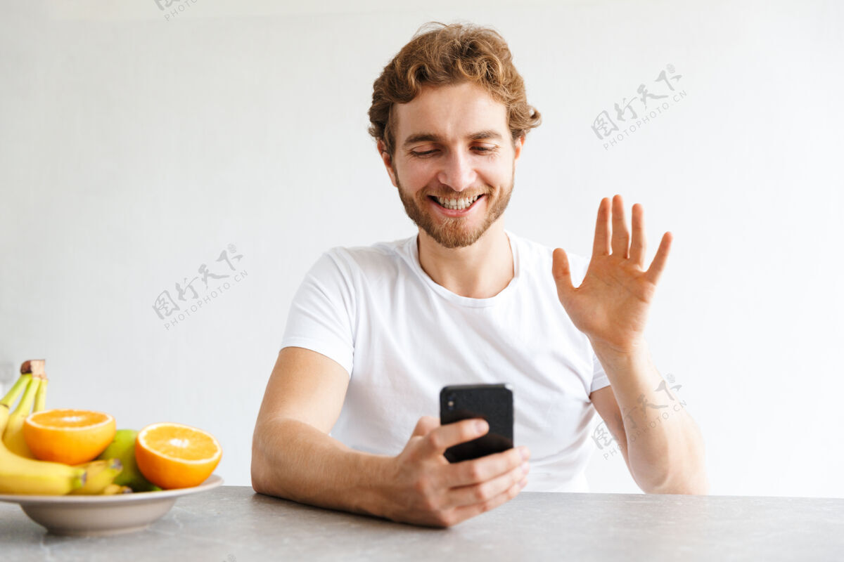 软件照片中 一个快乐的留着胡子的年轻人在家里的桌子旁用手机挥手交谈笔记本电脑通话休闲
