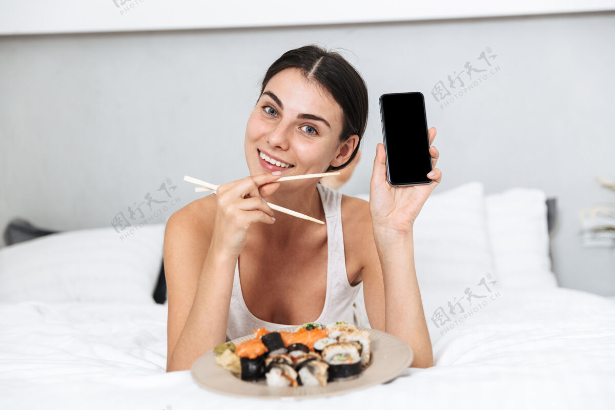 兴趣美丽的年轻女子在家里的床上放松 拿着手机 吃着盘子里的寿司放松卧室交流