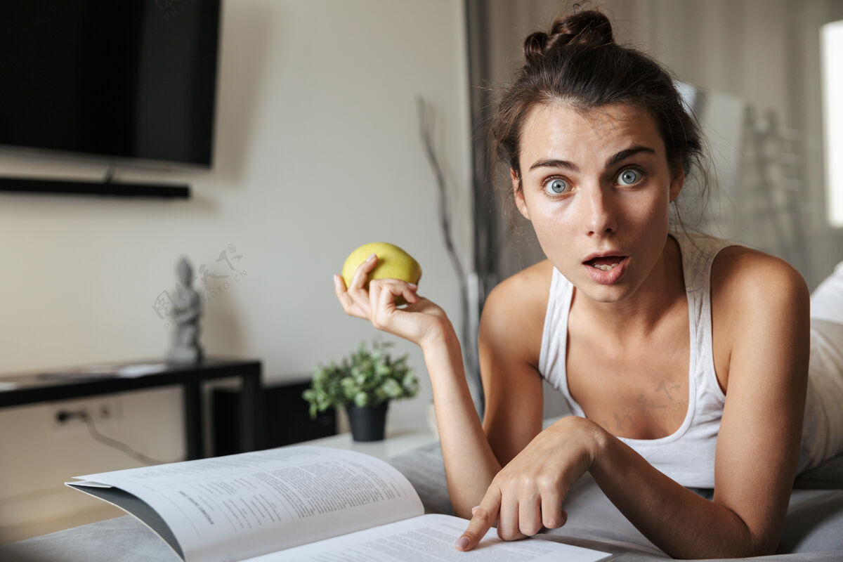 吸引力美丽的年轻女子在家的沙发上看书 吃青苹果苹果舒适学习