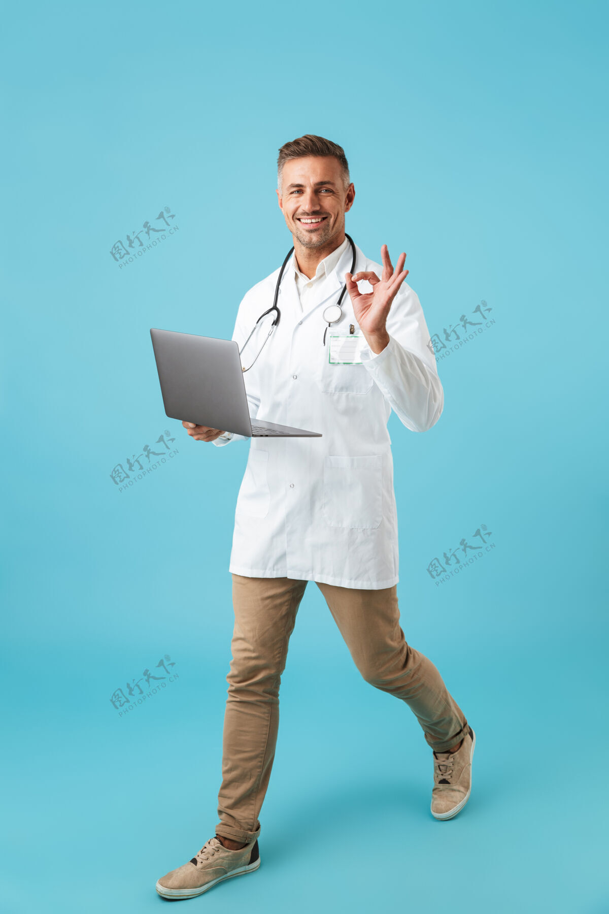 成人满脸笑容的男子身穿白色医用外套 手持听诊器 手持笔记本电脑行走 隔离在蓝色墙壁上笔记本电脑从业者职业