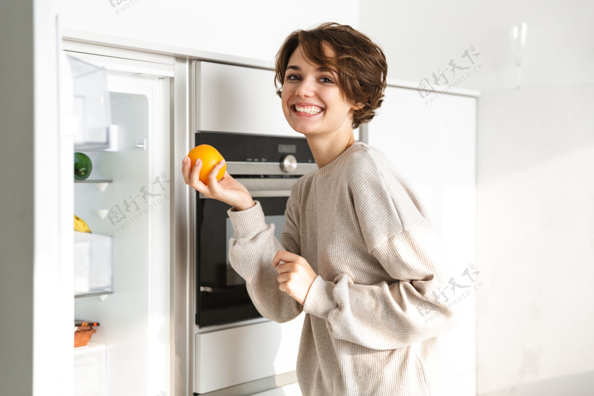 新鲜微笑的年轻女子站在冰箱前 手里拿着橘子概念膳食小吃