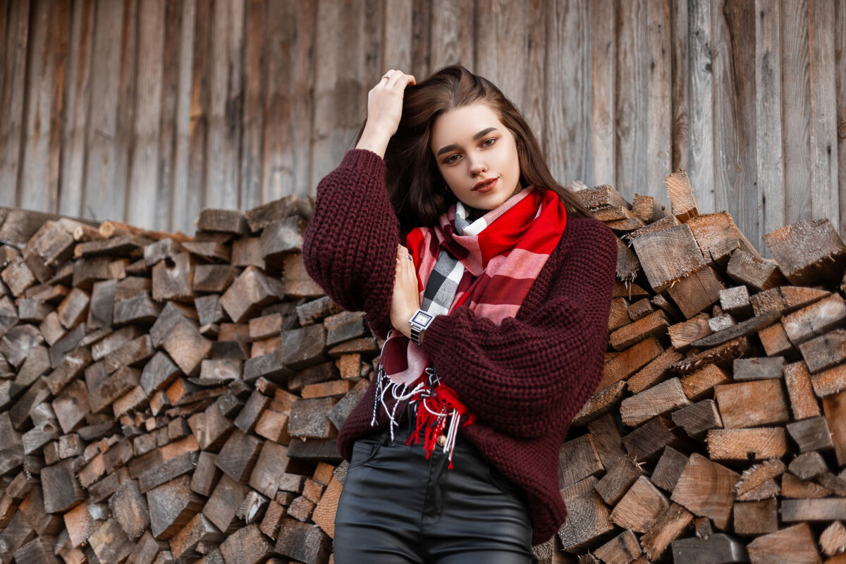 模特一个穿着针织毛衣 围着格子围巾 穿着皮裤的年轻女子在一个旧棚子附近摆姿势乡村棕色头发时尚