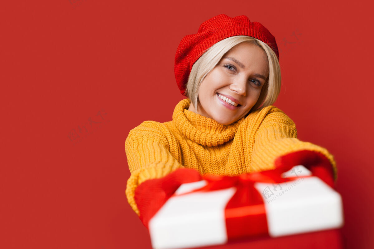 新年一位金发 头戴漂亮帽子的高加索女士微笑着向镜头赠送礼品盒 并在一个红色的工作室墙上做广告毛衣帽子礼物
