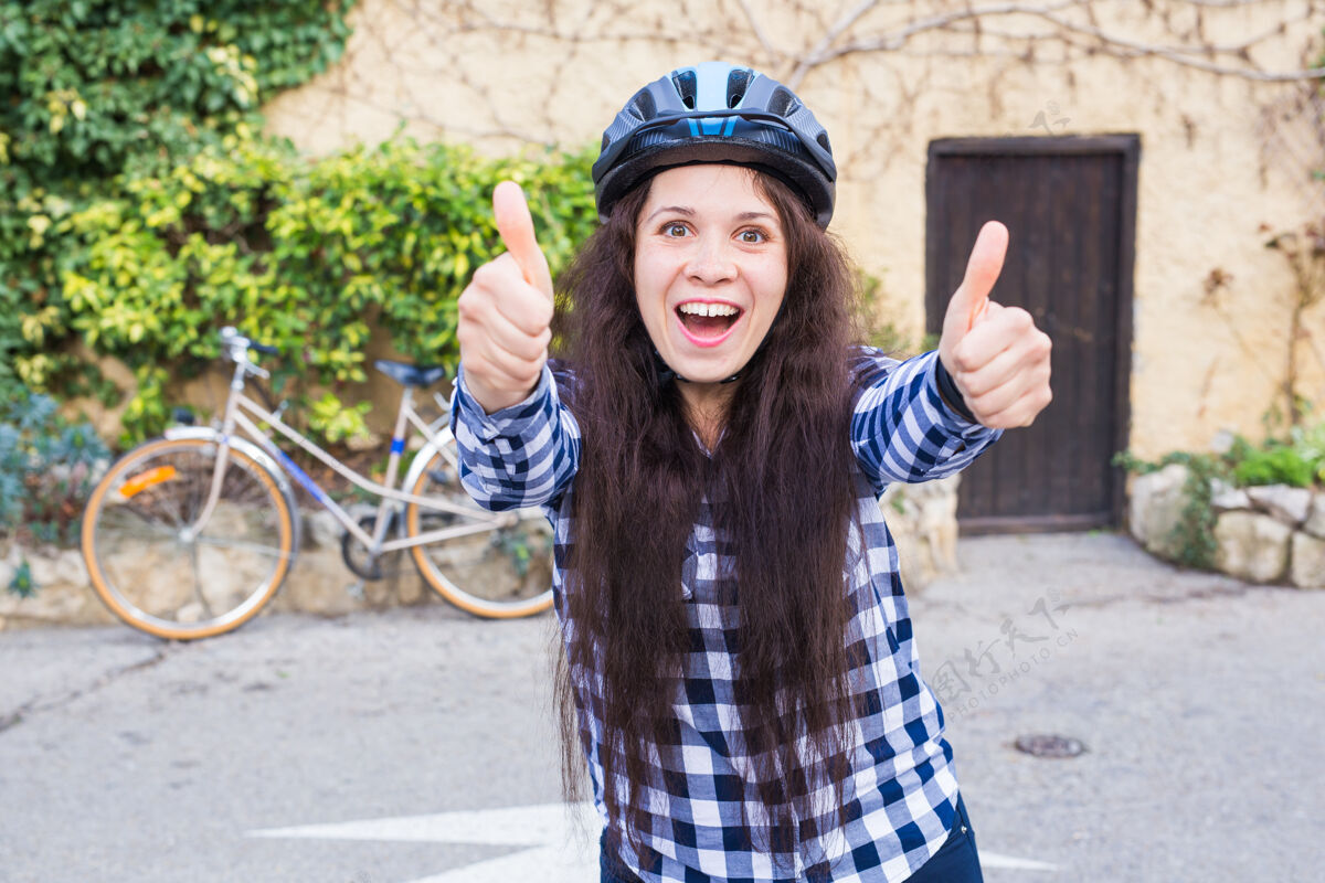 欢呼戴着头盔和太阳镜的微笑的年轻女子在墙上的小巷和自行车上竖起大拇指年轻人举行快乐