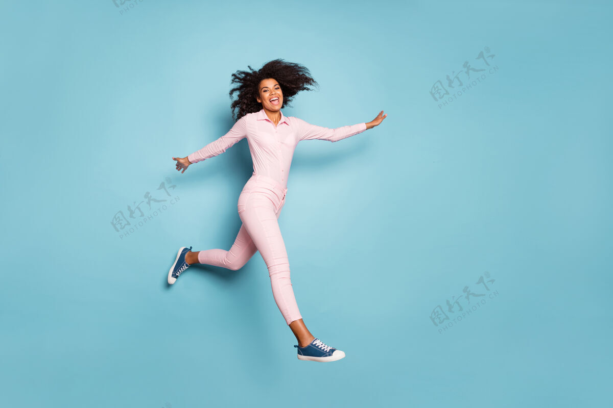 多民族令人惊异的深色皮肤的女士跳高展开双手的全长照片在外面度过最好的空闲时间穿粉红色衬衫裤子孤立的蓝色背景自由壁板运动