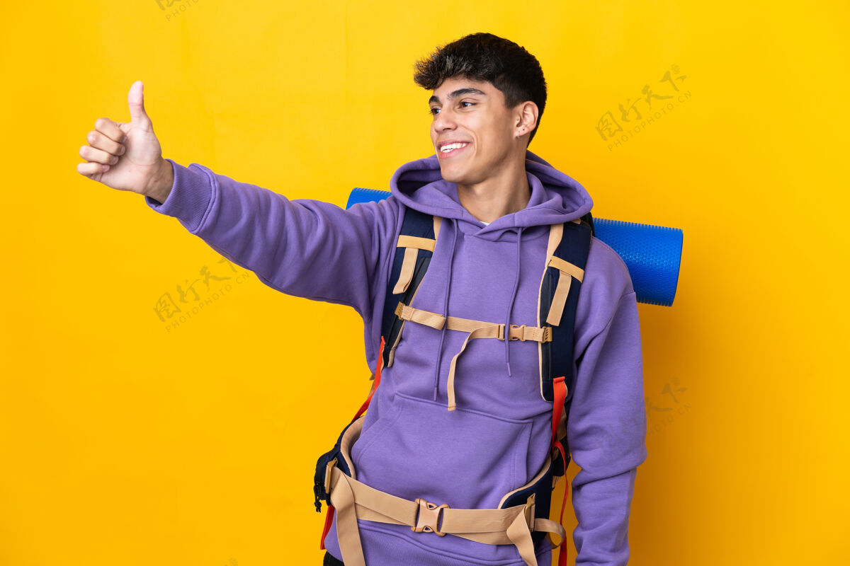 拇指一个年轻的登山者背着一个大背包 在孤立的黄色背景上竖起大拇指微笑好积极