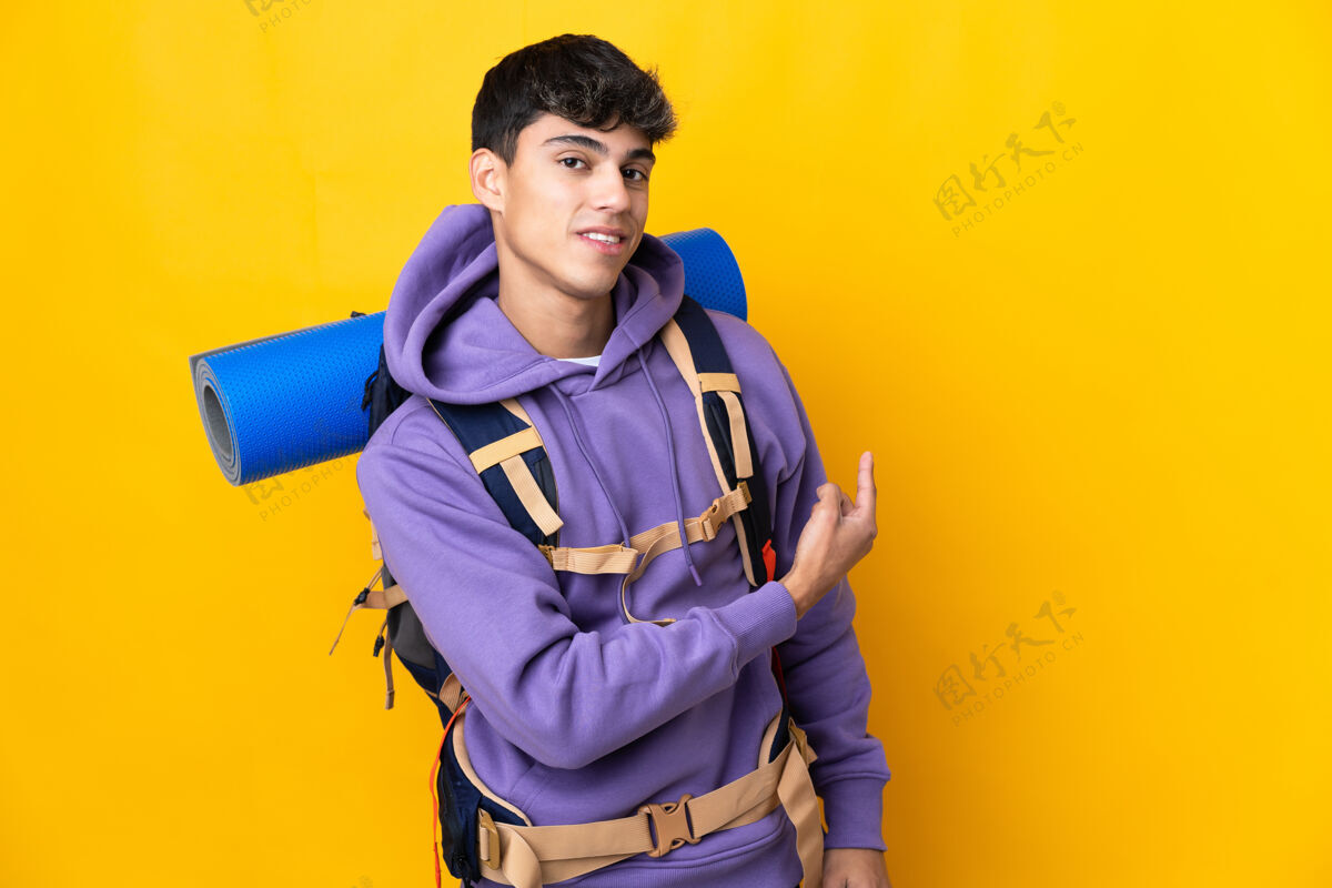 旅游一个年轻的登山者背着一个大背包 在孤立的黄色背景上指着后面年轻旅行好