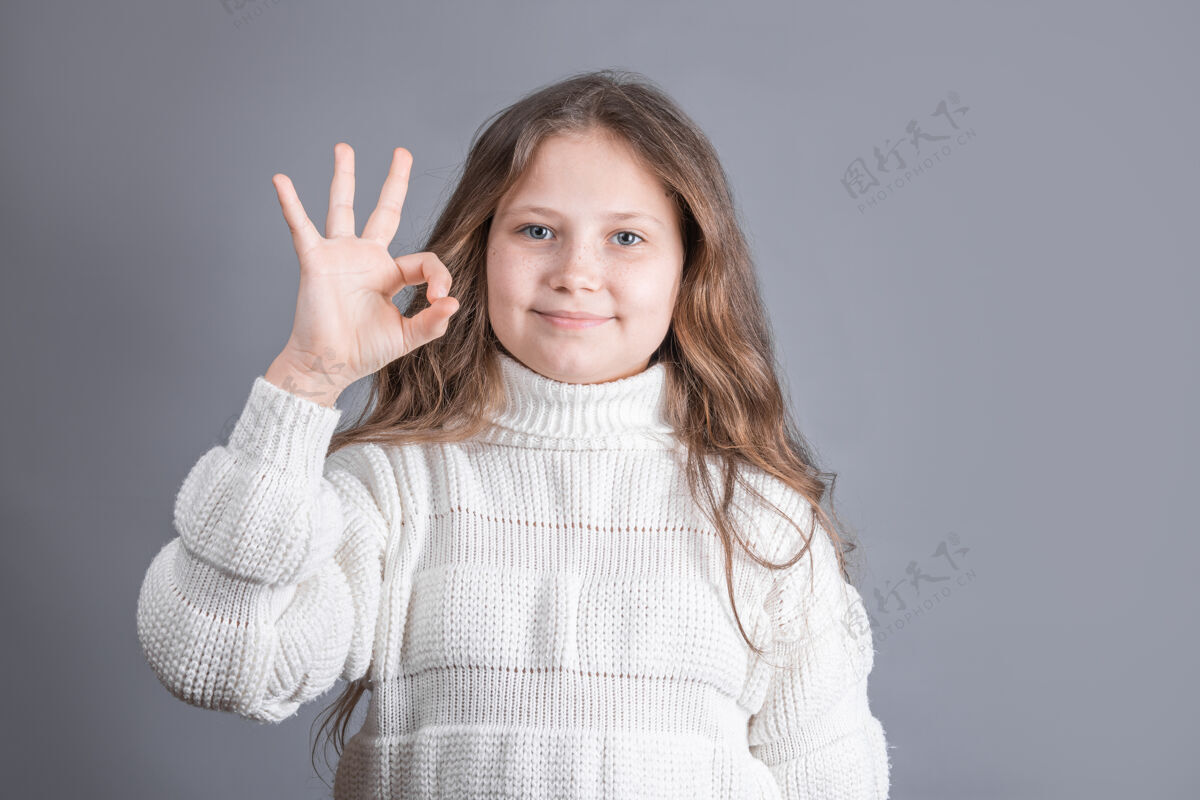 长一位金发 身穿白色毛衣 面带微笑的年轻迷人小女孩的肖像在一间灰色的工作室里展示了“OK”的标志背景.地点用于文本选择批准建议