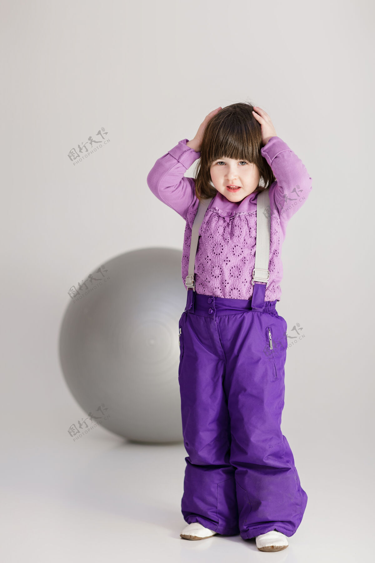 休闲穿着紫色衣服的可爱小女孩抱着他的头 在灰色背景上健身用的大球放松年轻大