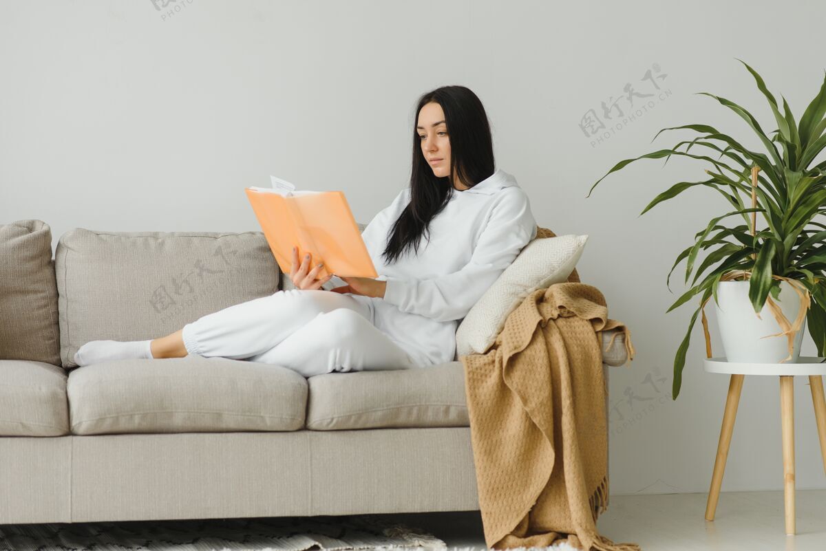 书漂亮的年轻女子躺在沙发上享受着在家看书的乐趣 穿着休闲装微笑着坐着阅读室内