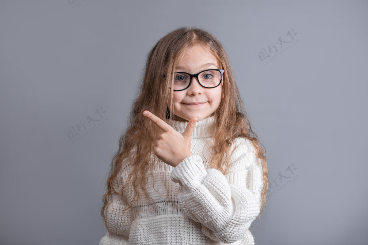 肖像在一个灰色的工作室里 一个年轻漂亮的小女孩的画像 金发飘逸 穿着白色毛衣 微笑着指着旁边背景.地点为了文本.复制空间时尚看小