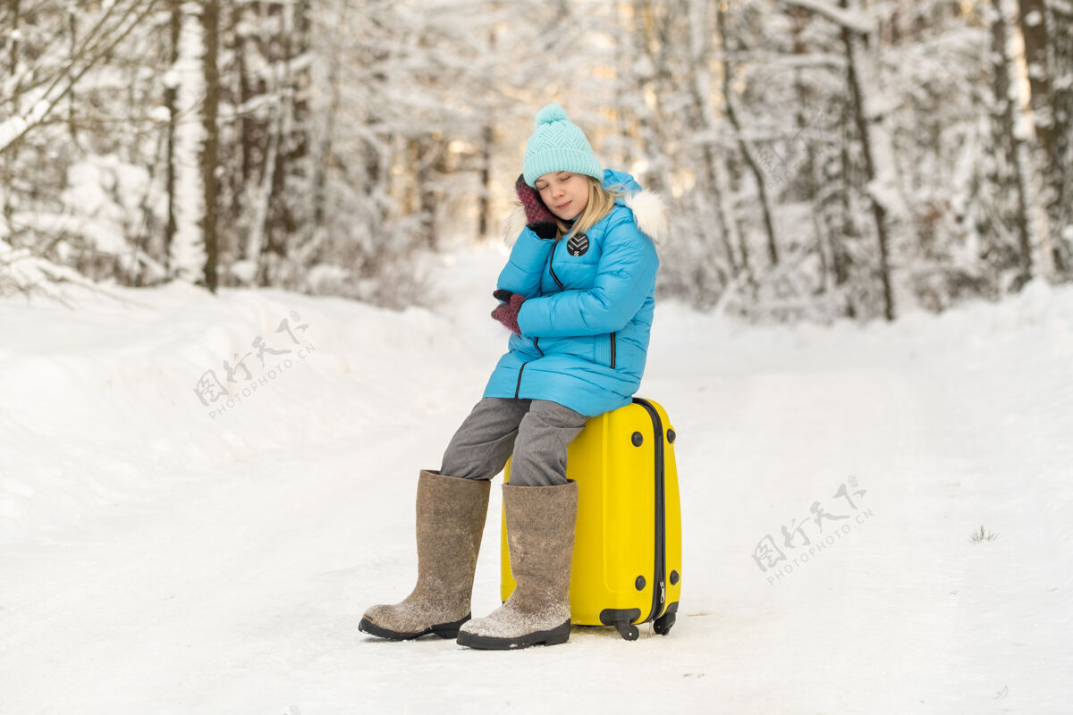 冒险冬天 一个穿着毡靴的女孩在一个寒冷的雪天坐在手提箱上霜冻郊区包
