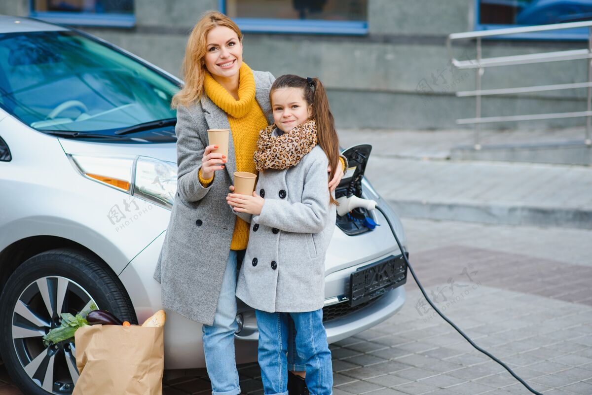 当代妈妈带着女儿在电动加油站给电动车充电 用手机说话互动母亲环境