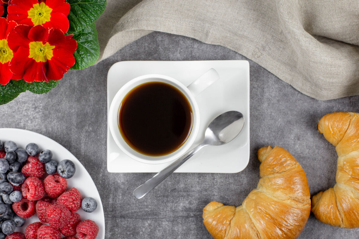 餐厅欧式早餐 羊角面包 白杯子里的黑咖啡 覆盆子 蓝莓和灰色水泥上的花水果花烘焙