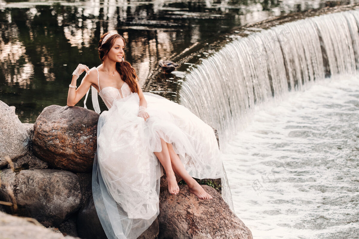 美丽一位穿着白色礼服 戴着手套 赤脚的优雅新娘正坐在公园的瀑布旁 享受着大自然浪漫女孩山河
